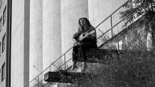 Low angle view of woman playing guitar while sitting against building