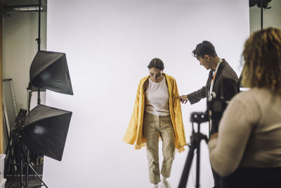 Male designer assisting female model against white backdrop during photo shoot in studio