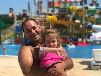 Portrait of happy shirtless father carrying daughter while standing at water park