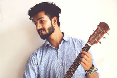 Young man playing guitar while standing against wall