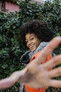 Smiling young woman gesturing while standing against green leaf wall