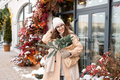 Stylish girl in glasses and hat near the shop window with christmas nobilis in the city
