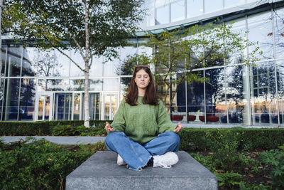 Young woman with eyes closed meditating on concrete seat in front of glass building