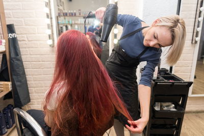 Female master dries long hair woman with a hair dryer in front a mirror in the salon hairdresser.