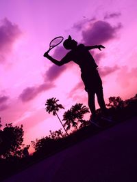 Tilt shot of silhouette man jumping while playing tennis against purple sky during sunset