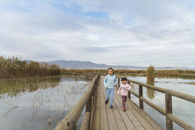 Scenic view of lake and two girl running on bridge against sky