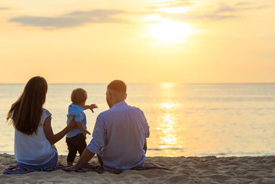 Rear view of family sitting at beach against sky during sunset