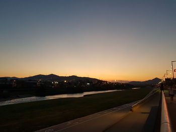 Road by illuminated city against clear sky during sunset