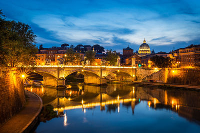 Bridge over river by illuminated buildings against sky at night