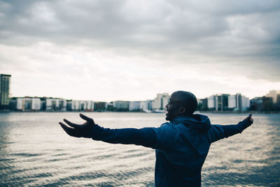 Male athlete standing with arms outstretched by sea against sky in city