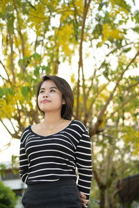 Low angle view of beautiful young woman looking away standing against trees in park