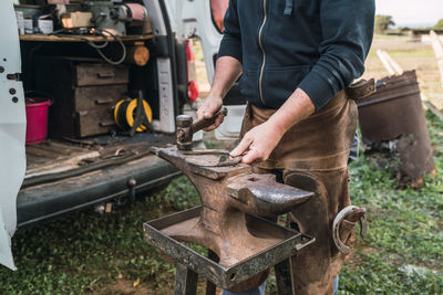 Crop faceless worker in uniform forging horseshoe with hammer on metal anvil while working in rural area on summer day