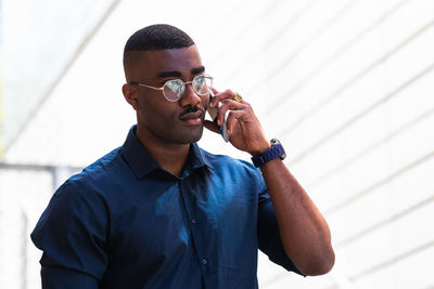 Confident african american adult guy in shirt and eyeglasses standing in city street while having conversation on mobile phone near stairs with railings in daylight