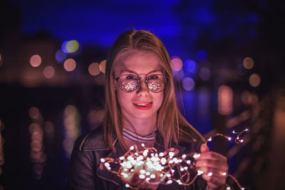 Portrait of young woman wearing eyeglasses holding illuminated string lights in city at night