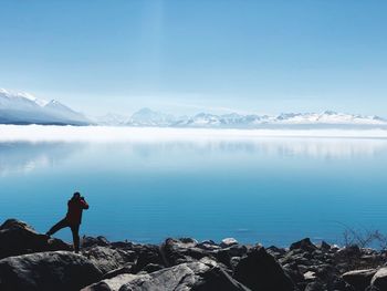 Rear view of man photographing while standing on rocks by lake