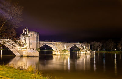 Bridge over river by illuminated buildings against sky at night