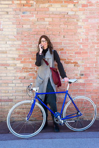 Smiling young woman with bike leaning on wall while using mobile