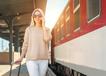 Blonde woman with her luggage go and speak in her mobile near red train rail station under sun light