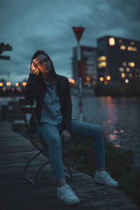 Portrait of young woman sitting on chair at riverbank in city during dusk