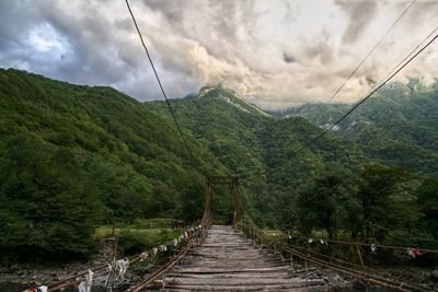 Footbridge amidst trees and mountains against sky