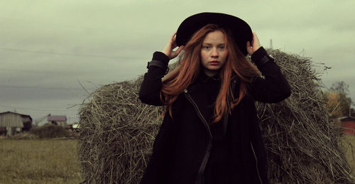 Portrait of young woman wearing hat while standing by hay bale against sky