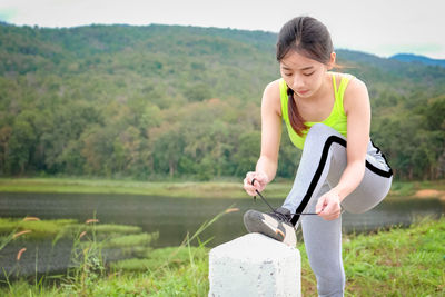 Young woman tying shoelace on stone by lake