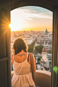 Rear view of woman standing at balcony against cityscape during sunset