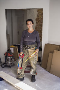 Portrait of confident female carpenter holding handsaw and ruler standing in apartment