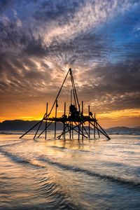 Silhouette oil pump on sea against sky during sunset