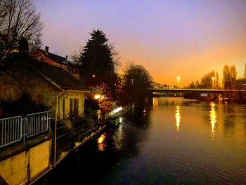 Illuminated buildings by river against sky at sunset