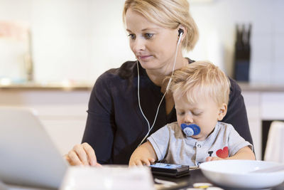 Baby boy using smart phone while mother listening music on laptop at table