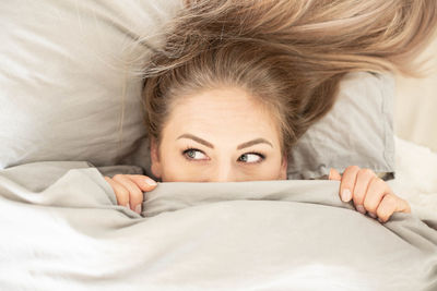 Directly above woman peeking from under blanket while lying on bed at home