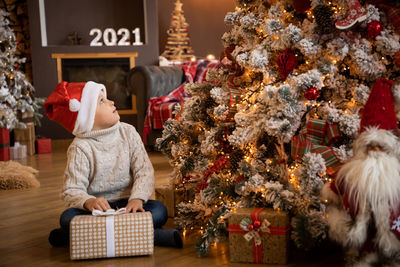 Cute boy holding gift box sitting by christmas tree at home