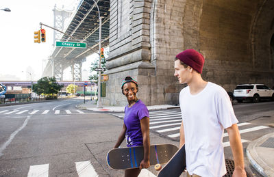 Young man with woman walking on road while holding skateboard