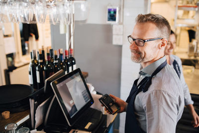 Smiling man using computer while holding credit card reader in restaurant