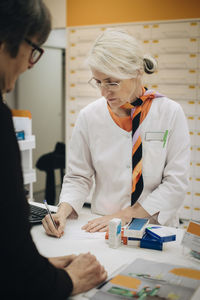 Mature female pharmacist writing on prescription paper at checkout with senior customer in medical store