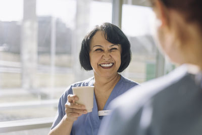 Happy female doctor holding disposable cup near colleague during coffee break at hospital