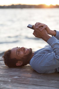 Happy man using mobile phone while lying on wooden pier against sea during sunset