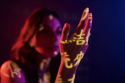 Fashionable young female model with light projection in shape of oriental hieroglyphs looking at extended hand in dark studio with red illumination