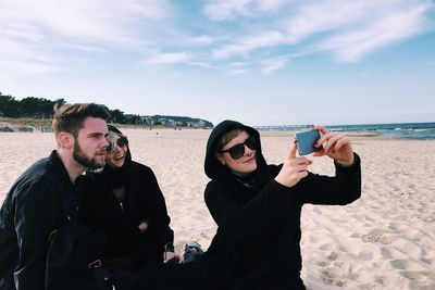 Young friends taking selfie at beach against sky