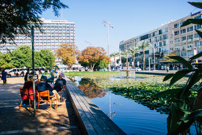 People sitting by canal in city against sky