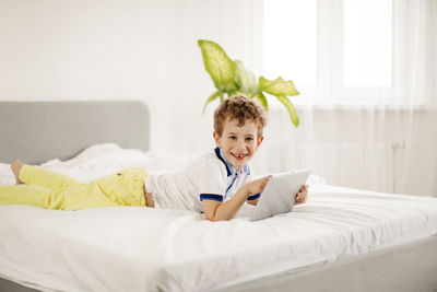 Curly-haired boy is lying on the bed in the bedroom and playing computer games on a tablet