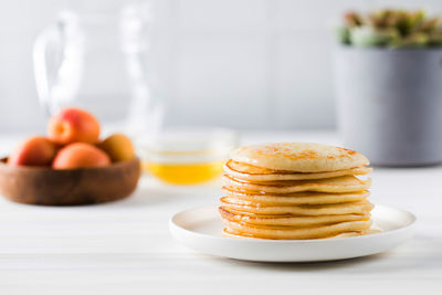 The pancakes are stacked in a plate. in the background, a bowl of honey, fruit, and a jug of milk.