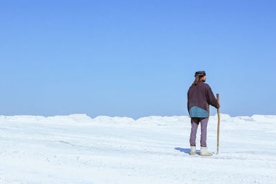 Back view of a man in turban walking with a cane among the white plain under a clear blue sky.