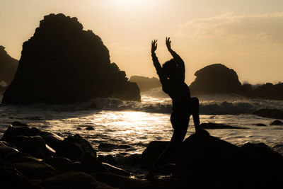 Silhouette man on rock at beach against sky during sunset