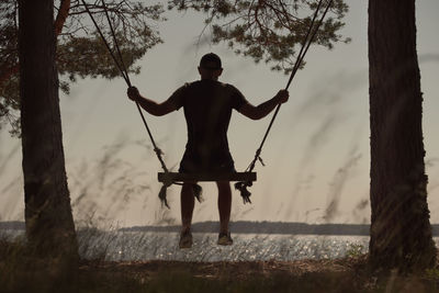 A young man swings on a children's swing by the lake. back view. themes of childhood and memories