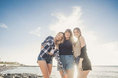 Portrait of young friends standing on shore at beach against sky on sunny day