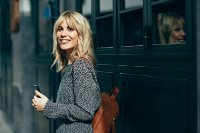 Portrait of young woman holding smart phone while standing against building