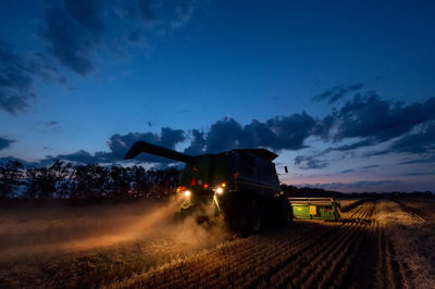 Illuminated combine harvester on agricultural field against sky at dusk