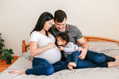 Family asian chinese pregnant woman and caucasian man with toddler girl sitting on bed at home. 